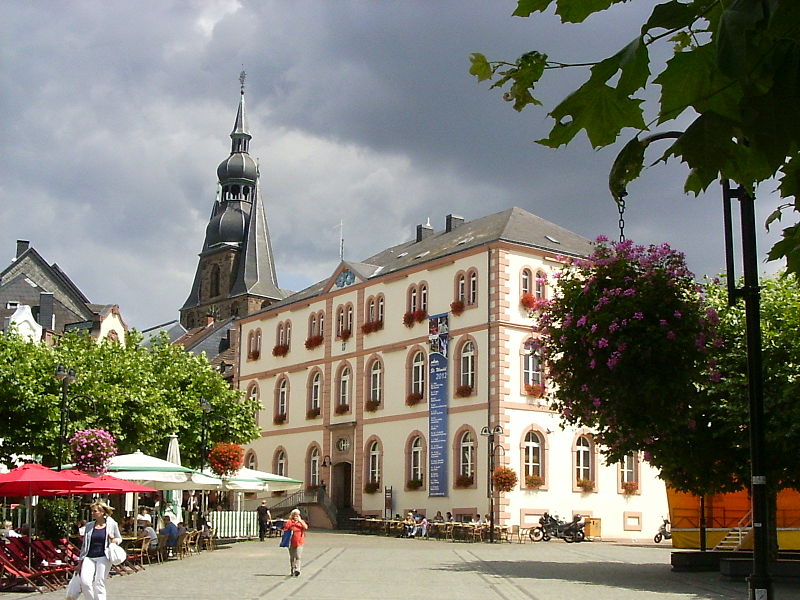 Blick auf den Schlossplatz St. Wendel, einen von Bäumen umsäumten Platz mit Außengastronomie. Im Hintergrund sind das alte Rathaus und die Wendelsbasilika zu sehen.