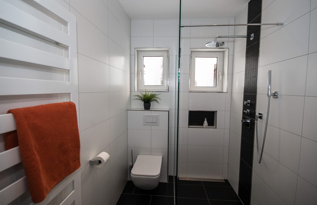 Blick ins gefließte Badezimmer mit begehbarer Dusche inklusive Regenbrause, Fußbodenheizung, Toilette.