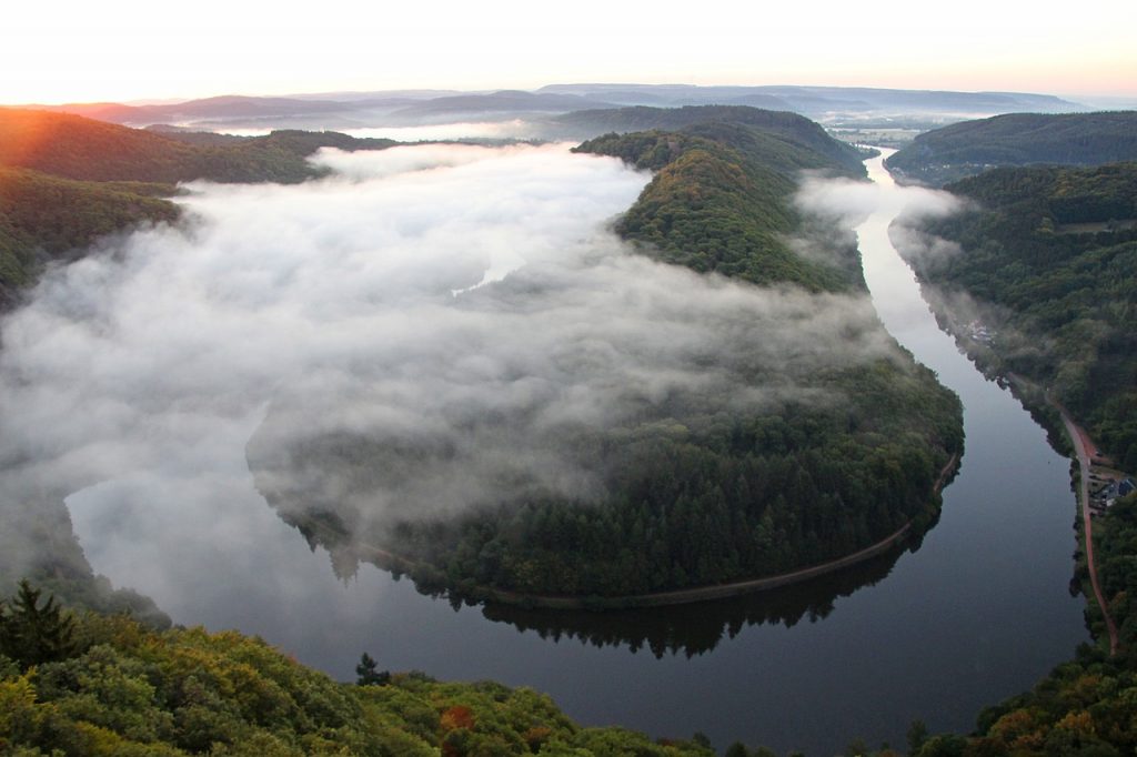 Blick auf die Saarschleife von Oben, einen Flusslauf, welcher sich U-förmig durch waldige Landschaft schlängelt. Über dem Fluss liegt Nebel, am linken Rand geht die Sonne auf.